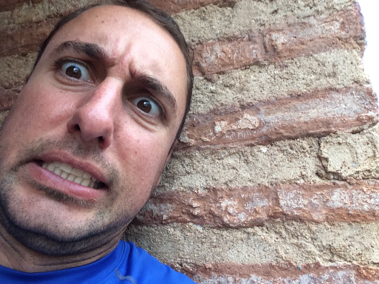 A man in a blue shirt stands in front of a brick wall with a confused/concerned look on his face.