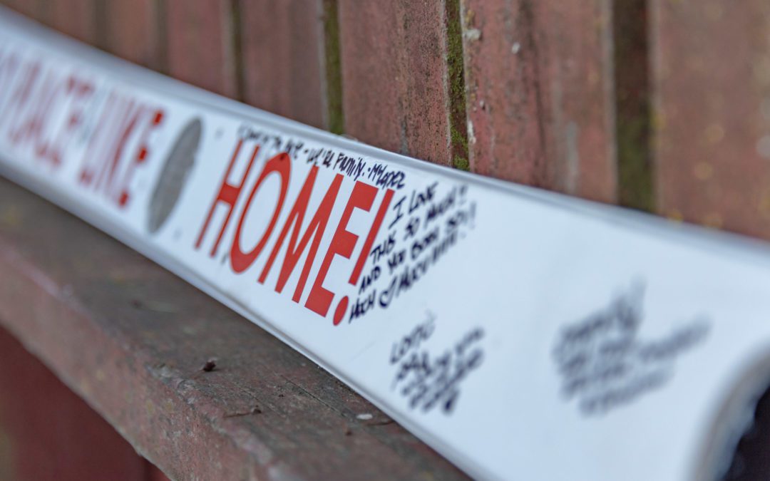 A white shot ski with the word "Home!" printed in red sits on a dull fence.