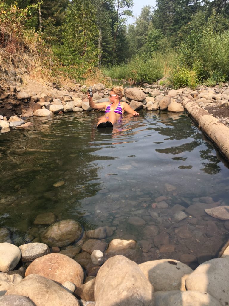 A woman wearing a purple striped bathing suit sits at the end of a hot spring pool enjoys a beer while looking off into the distance.