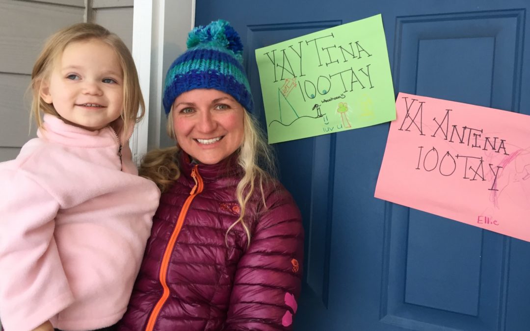 A woman wearing a pink jacket and blue striped hat stands holding a two year old girl in a pink fleece. They're in front of a door with two signs, one that reads "YAY Tina 100TAY" and the other "TA Antina 100 TAY"