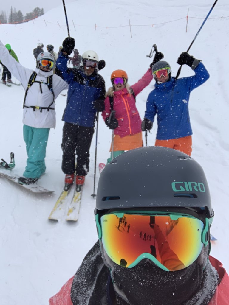 A person wearing a ski helmet and goggles takes a selfie in front of four other excited skiers in a whiteout.