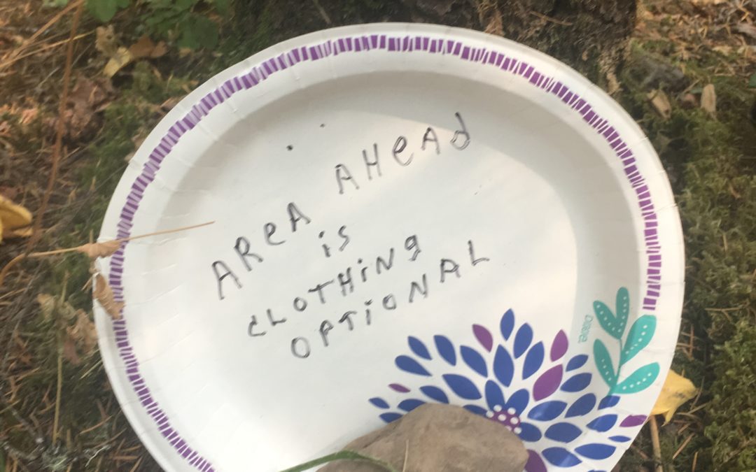 A white paper plate with "Area Ahead Is Clothing Optional" handwritten in the middle, sits leaning against a tree on a forest floor.