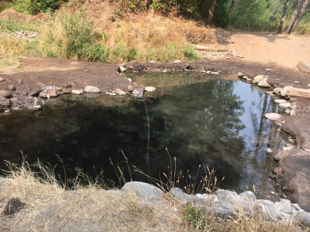 A clear blue pool in the shape of a heart sits surrounded by brown dirt, with trees on a hillside in teh distance.