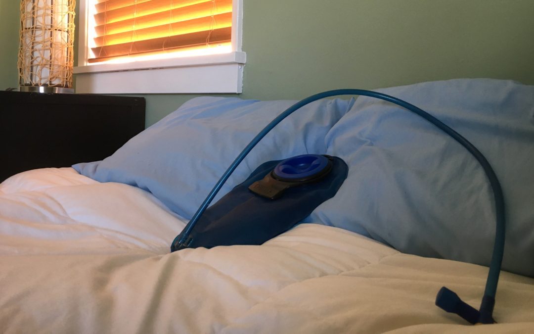 A full camelback bladder sits on a white sheet and blue pillows of a bed.