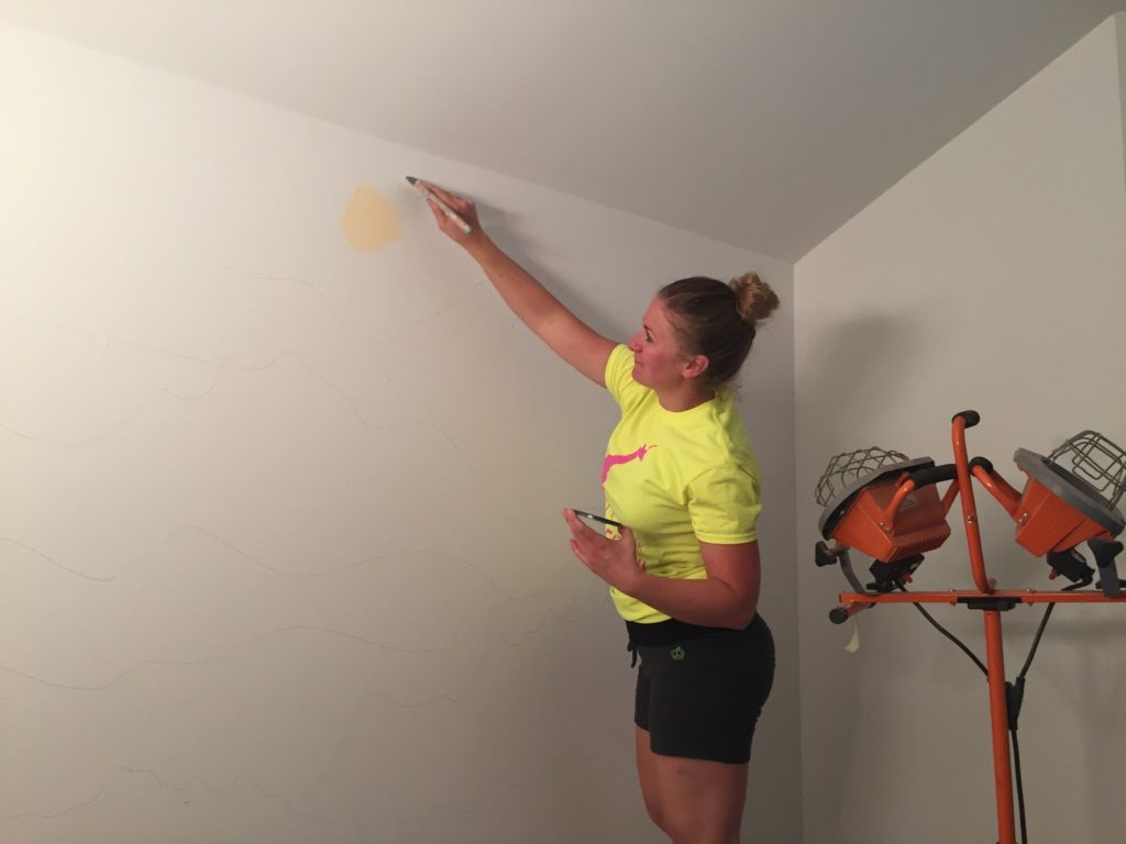 A woman in a yellow shirt and grey shorts puts the first color of peach paint on a blank white wall.