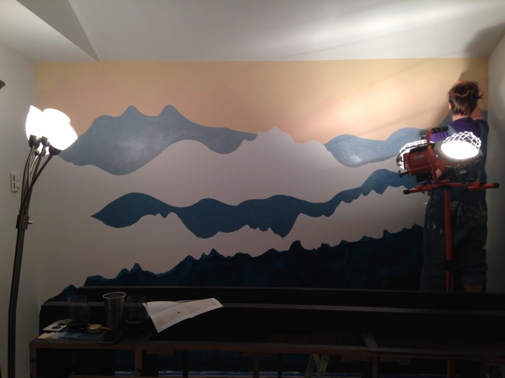 A woman adds layers of paint to a bedroom mountain mural.