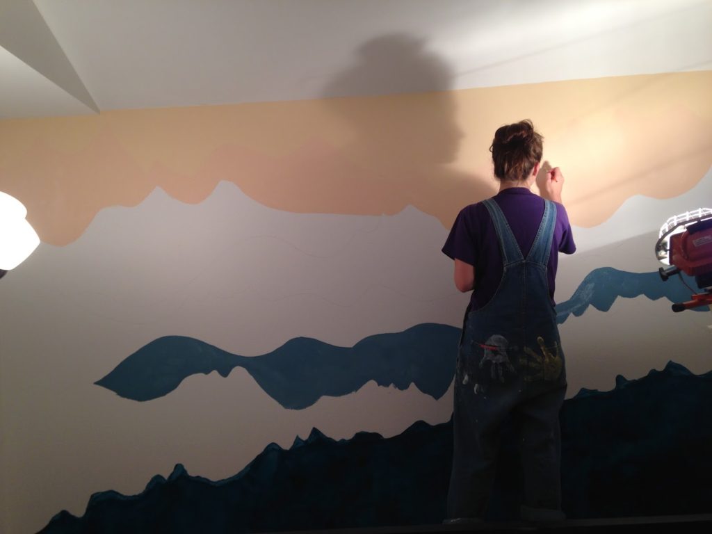 A woman in blue overalls covered in paint stands in front of an incomplete wall mural of a mountain.