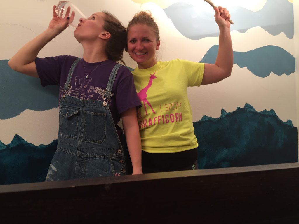 Two women standing in front of an incomplete mountain mural painting take a break to drink wine.