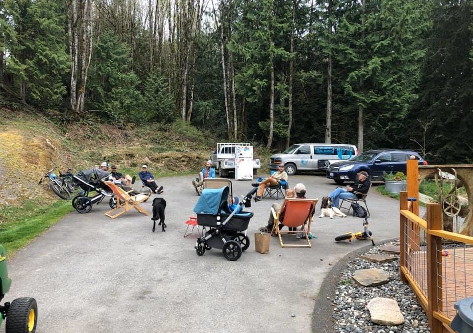 Seven people sit on a large concrete pad at the end of a driveway in the woods 6-feet apart from each other. A kegorator sits in the back. COVID made them do it.