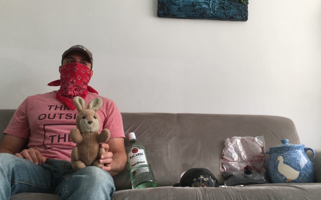 A man wearing a bandana holds a stuffed bunny while sitting on a couch next to a handle of bacardi, a large rock, a bag of wine, and an ugly vase. These are all items you can use to practice crevasse rescue.