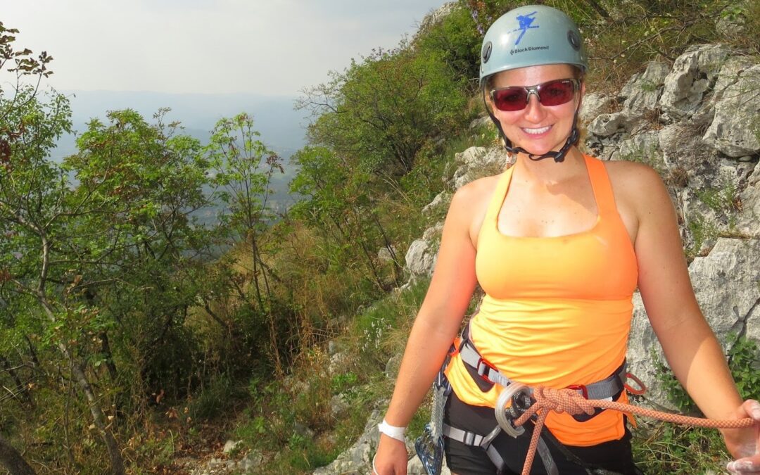 A white woman in an orange tanktop stands in front of a tropical landscape wearing a blue helmet and a grey climbing harness tied into an organ climbing rope.